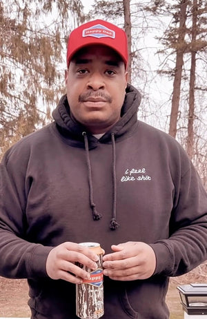 Man wearing a red cap and dark hoodie holding a can outdoors with trees in the background